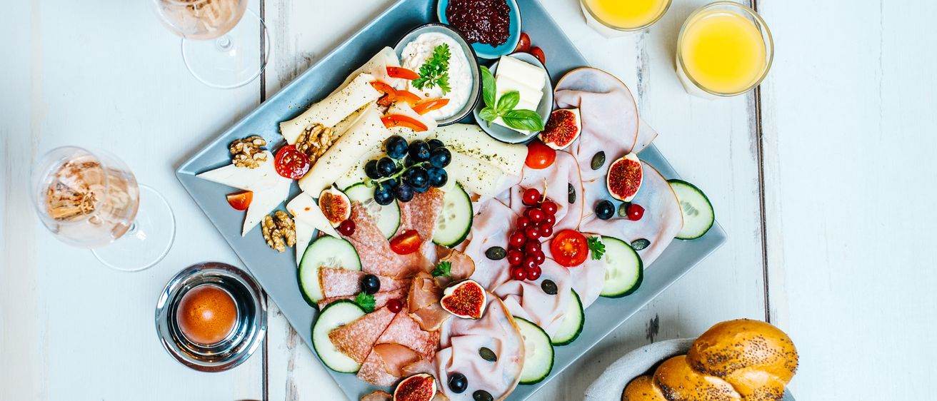 Ein blaues Tablett mit verschiedenen Lebensmitteln wie Käse, Früchten, Feigen und Fleisch, zusammen mit zwei Gläsern Orangensaft, zwei Weingläsern und einer Schüssel mit Brot.
