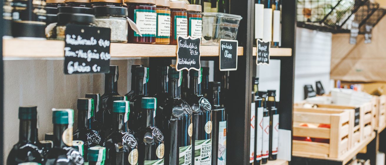 Ein Regal gefüllt mit verschiedenen Flaschen Olivenöl und Gläsern mit Konserven, jeweils mit ihren Inhalten und Preisen beschriftet. Im Hintergrund befinden sich Holzlagerboxen.