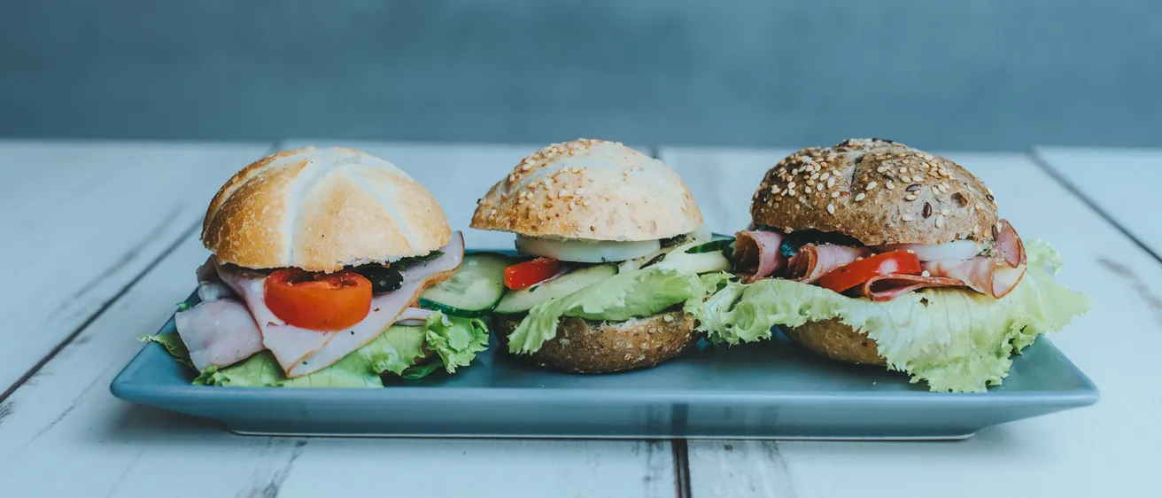 Drei Sandwiches mit verschiedenen Füllungen, einschließlich Salat, Tomaten und Gurken, sind ordentlich auf einem blauen Tablett auf einem weißen Tisch angeordnet.