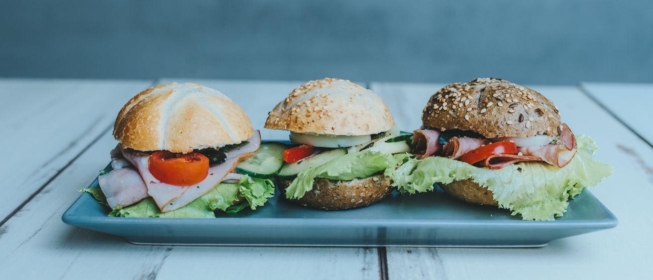 Drei Sandwiches mit verschiedenen Füllungen, einschließlich Salat, Tomaten und Gurken, sind ordentlich auf einem blauen Tablett auf einem weißen Tisch angeordnet.