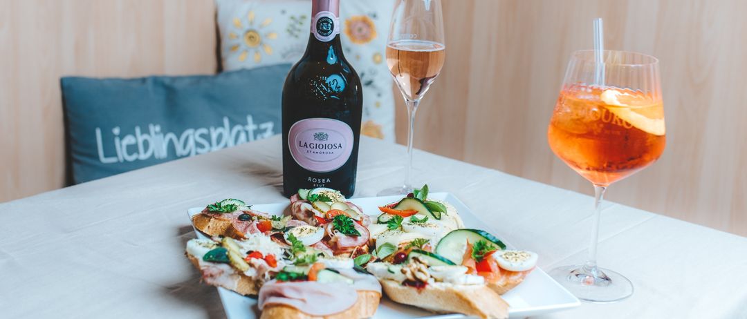 Eine Flasche Lagioiosa Rosea steht neben zwei Getränkegläsern auf einem Tisch, begleitet von einem Teller mit Sandwiches mit verschiedenen Belägen.