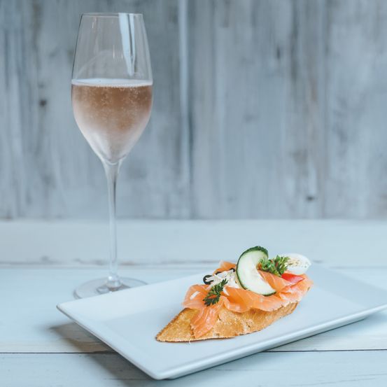 Ein Glas Champagner steht neben einem weißen Teller, der ein offenes Sandwich mit Räucherlachs, Gurke und Eierscheiben auf einem hölzernen Hintergrund enthält.