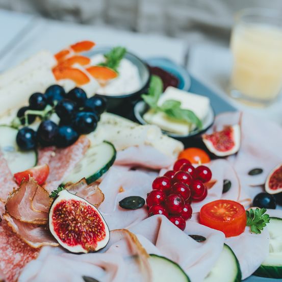 Ein Nahaufnahme einer Charcuterie-Platte mit Feigen, Trauben, Tomaten und geschnittenem Fleisch, begleitet von einem Glas Saft.