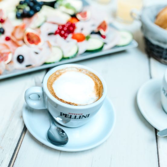 Eine Tasse Espresso mit Schaum auf einer Untertasse auf einem Tisch mit einem Tablett mit Obst und Brot im Hintergrund.