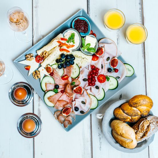 Ein blaues Tablett enthält eine Auswahl an Fleisch, Käse, Obst und Gemüse. Daneben stehen drei Gläser mit Saft und zwei Weingläser auf einem weißen Tisch.