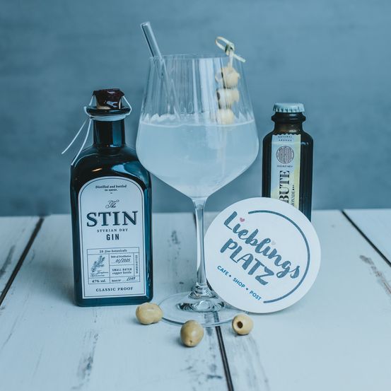 Ein Glas Gin mit Strohhalm, eine Flasche Gin und eine kleine Flasche Likör auf einem Tisch mit einem Untersetzer.