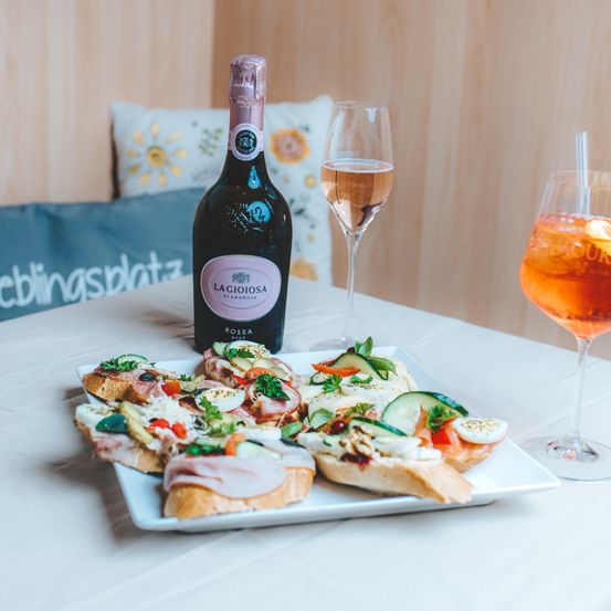 Eine Flasche La Gioiosa Rosea steht neben zwei Gläsern auf einem Tisch mit einer Platte verschiedener Sandwiches, garniert mit Gemüse und Fleisch.