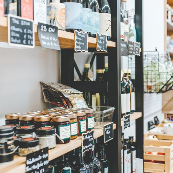 Ein Geschäft mit Regalen, die verschiedene Lebensmittel präsentieren. Flaschen Wein, Marmeladen und andere Artikel sind ordentlich angeordnet. Etiketten und Preisschilder sind an den Artikeln sichtbar.
