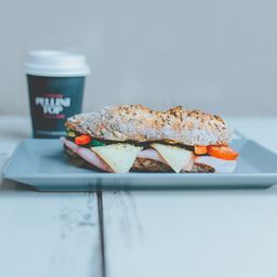 Ein Sandwich mit Schinken, Käse und Tomatenscheiben steht auf einem Teller mit einem Kaffeebecher. Ein roter Apfel ist links.