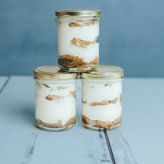 Drei Glasgläser mit goldenen Deckeln enthalten ein cremiges Dessert mit Keksboden, vor einem blauen Hintergrund präsentiert.