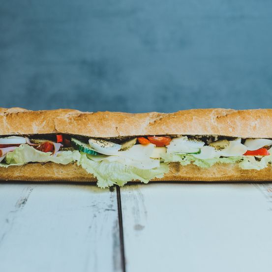 Ein Sandwich gefüllt mit Salat, Tomaten, Gurken, Ei und Käse auf einem weißen Holztisch mit blauem Hintergrund.