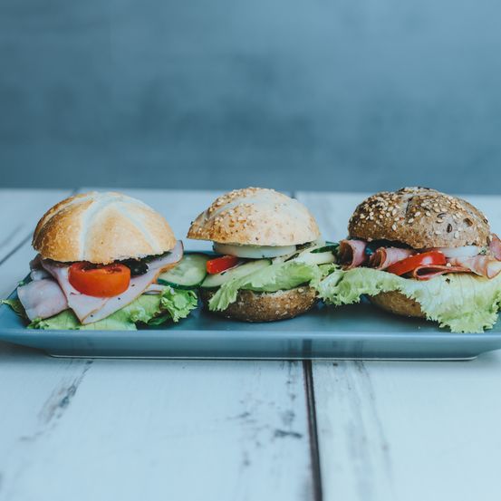 Drei Sandwiches mit Sesambrötchen sind auf einem blauen Teller. Sie enthalten Schinken, Tomate, Gurke und Salat. Der Hintergrund ist eine hellblaue Wand.