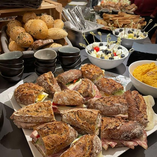 Ein Tisch mit verschiedenen Speisen, darunter Sandwiches, Hummus, Brot und Vorspeisen. Schüsseln mit Oliven und ein Charcuterie-Platte sind ebenfalls vorhanden.