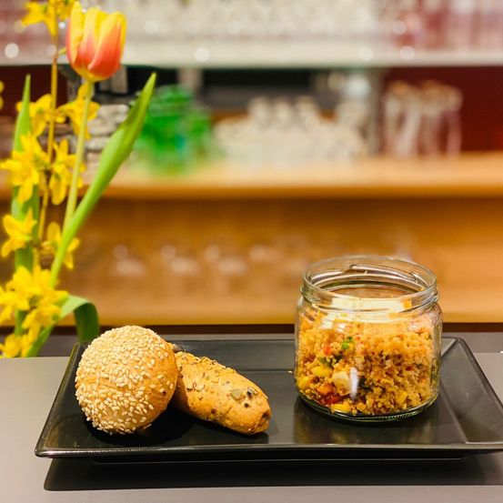 Ein schwarzer Teller enthält ein Sesambrötchen, ein Brötchen und ein Glas Quinoa-Salat. Eine gelbe Blume im Hintergrund.