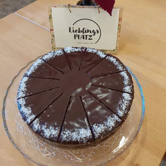 Ein Schokoladenkuchen auf einem Teller, in Stücke geschnitten, mit einem Schild im Hintergrund, das 'Lieblings Platz' steht. Der Kuchen ist mit Schokolade und weißen Streuseln verziert.