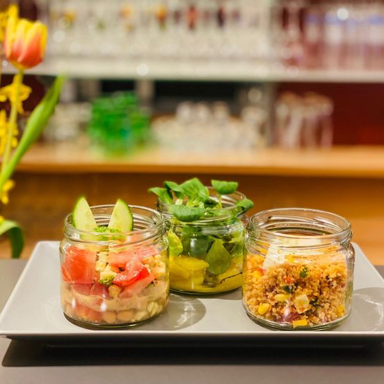 Drei Glasgläser mit verschiedenen Salaten, eines mit Tomaten, eines mit grünem Gemüse und eines mit Getreide, auf einem weißen Teller.