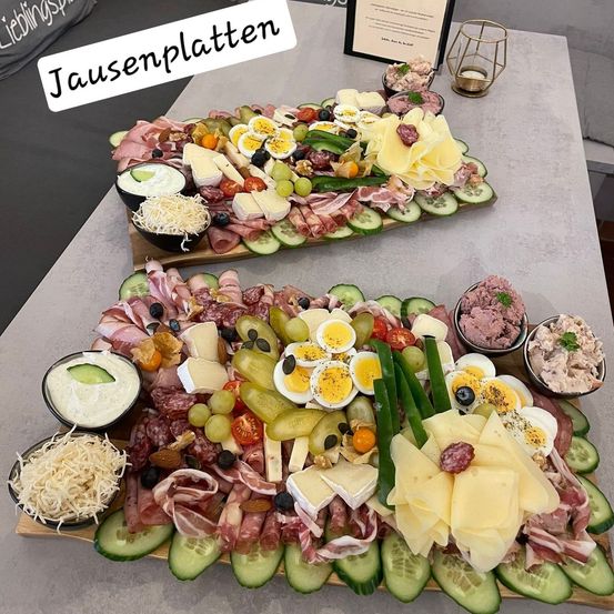 Zwei große Charcuterie-Bretter mit verschiedenen Fleischsorten, Käse, Gemüse und Dips stehen auf einem Tisch. Ein Schild mit dem Text 'Lieblingsplatz' befindet sich hinter den Brettern. Links sind Kissen mit dem Text 'Lieblingsplatz'. Rechts steht ein Kerzenhalter.