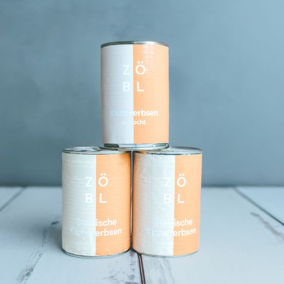 Drei Dosen Zölbl Küchenerbsen stehen gestapelt auf einer Holzoberfläche. Jede Dose hat ein weiß-oranges Etikett mit Markenname und Produkttyp.
