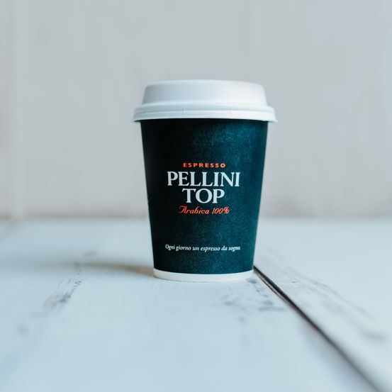 Eine schwarze Kaffeetasse mit weißem Deckel auf einem Holztisch. Sie ist mit 'ESPRESSO PELLINI TOP Arabica 100%' beschriftet.