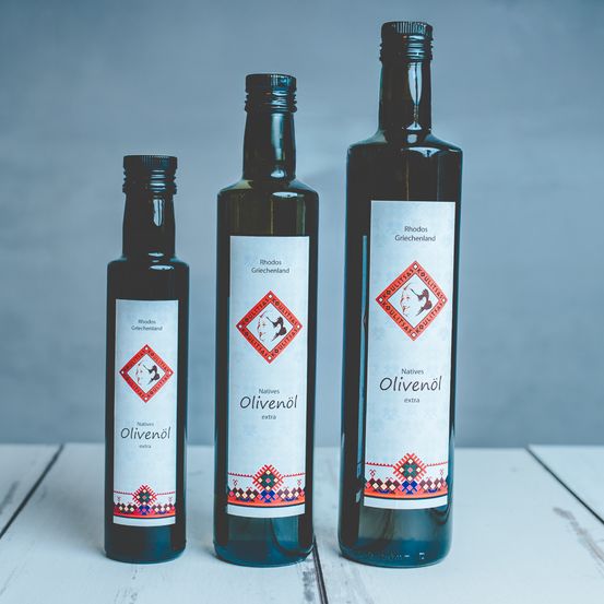 Drei Flaschen Olivenöl mit der Aufschrift 'Natives Olivenöl extra' stehen auf einem Holztisch vor einem grauen Hintergrund.