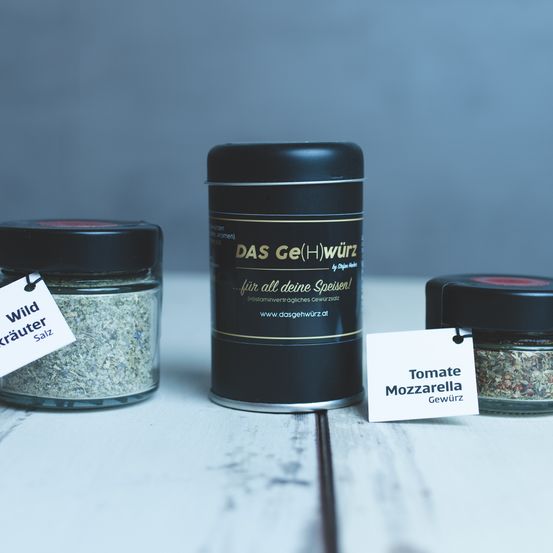 Drei Gläser mit Gewürzen stehen auf einem Holztisch. Das mittlere Glas ist mit 'DAS Ge(H)wurz' beschriftet. Das linke Glas hat ein Etikett für 'Wild Krauter Salz'. Das rechte Glas hat ein Etikett für 'Tomate Mozzarella Gewürz'.