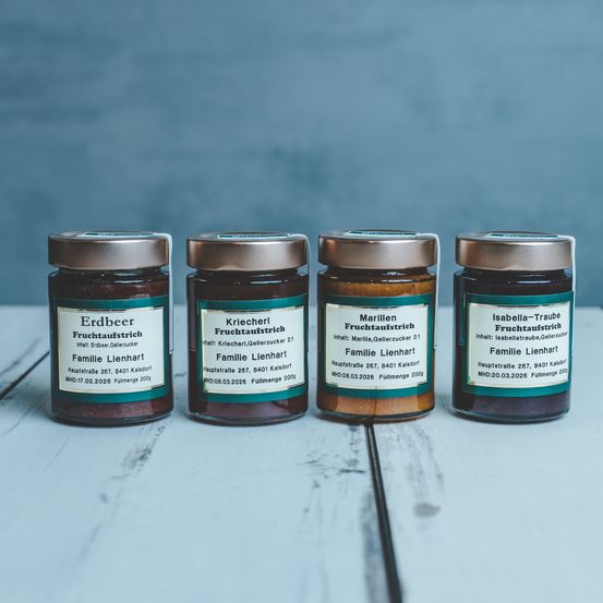 Vier Gläser mit Konfitüren mit Etiketten: Erdbeere Fruchtaufstrich, Kriecher Fruchtaufstrich, Marille Fruchtaufstrich, Isabella-Traube Fruchtaufstrich. Jedes Glas hat einen goldenen Deckel. Auf einer Holzoberfläche.