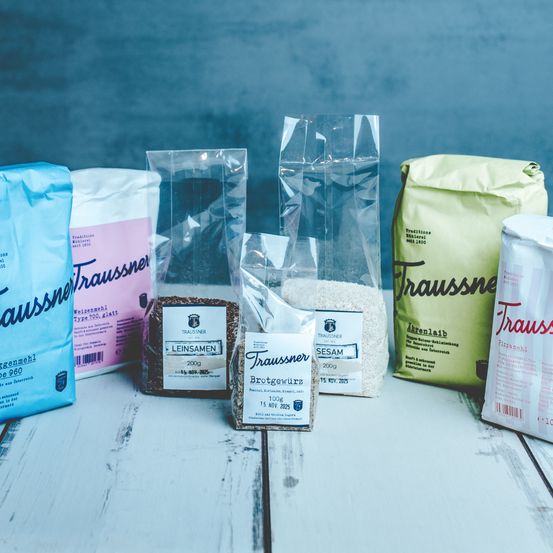 Verschiedene Mehl-, Getreide- und Samenbeutel sind auf einem Holztisch angeordnet, darunter Beutel mit Leinsamen, Brotgewürz und Sesam.