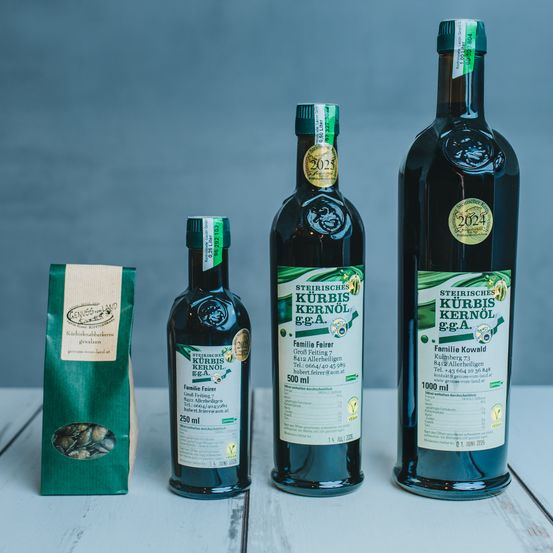 Drei Flaschen des grün gekennzeichneten 'Steirisches Kurbis Kernol' sind ausgestellt. Die Flaschen variieren in der Größe, wobei eine mit 'Familii Feier' 250 ml und eine andere mit 'Familii Kowald' 1000 ml gekennzeichnet ist. Ein grüner Beutel mit Samen ist ebenfalls vorhanden.