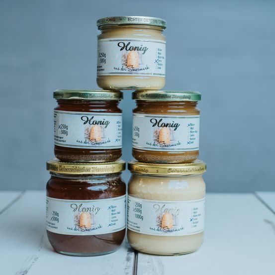 Vier Gläser mit Honig sind auf einer weißen Oberfläche gestapelt. Jedes Glas hat einen anderen Geschmack und ist mit seinem Gewicht und Herkunft gekennzeichnet. Die Gläser haben goldene Deckel und weiße Etiketten mit einer Zeichnung eines Honigwaben.
