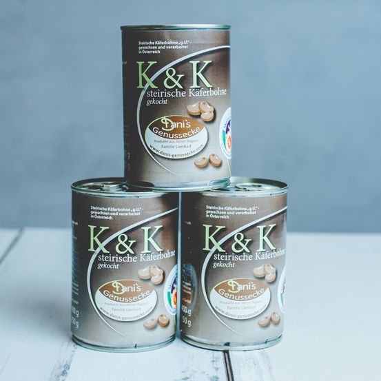 Drei Dosen K&K Steirische Kaferbohne, ein in Österreich angebautes und verarbeitetes Produkt, sind auf einer Holzoberfläche gestapelt. Die Dosen haben braune und grüne Etiketten mit dem Markennamen und den Produktdetails.