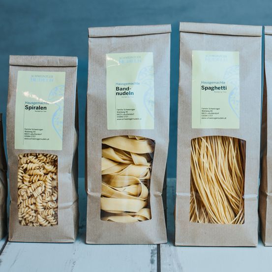 Vier braune Papiertüten mit Etiketten sind zu sehen. Die erste Tüte hat ein Etikett mit der Aufschrift 'Hausgemachte Spiralen' und einem Bild von Spiralnudeln. Die zweite Tüte sagt 'Hausgemachte Bandnudeln' mit einem Bild von Bandnudeln. Die dritte Tüte sagt 'Hausgemachte Spaghetti' mit einem Bild von Spaghetti. Die vierte Tüte ist ebenfalls mit 'Hausgemachte Spaghetti' beschriftet. Jede Tüte ist geöffnet und zeigt die Nudeln darin.
