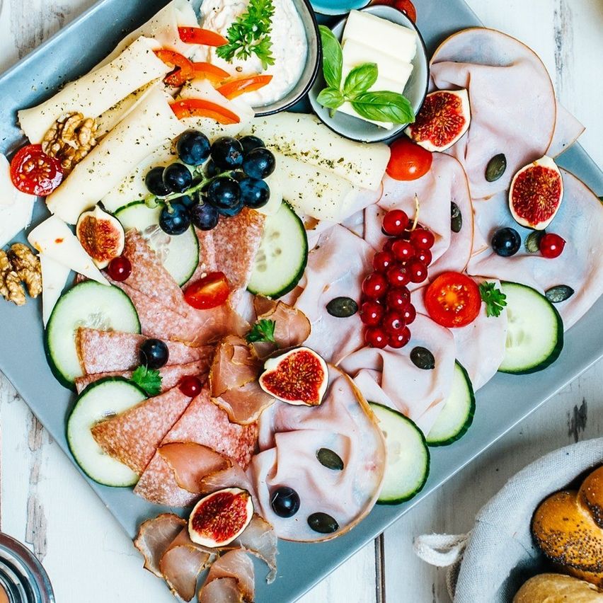 Ein blauer Teller mit verschiedenen Lebensmitteln wie Fleisch, Käse, Obst und Gemüse, begleitet von Brot, Eiern und Gläsern Orangensaft auf einem weißen Tisch.
