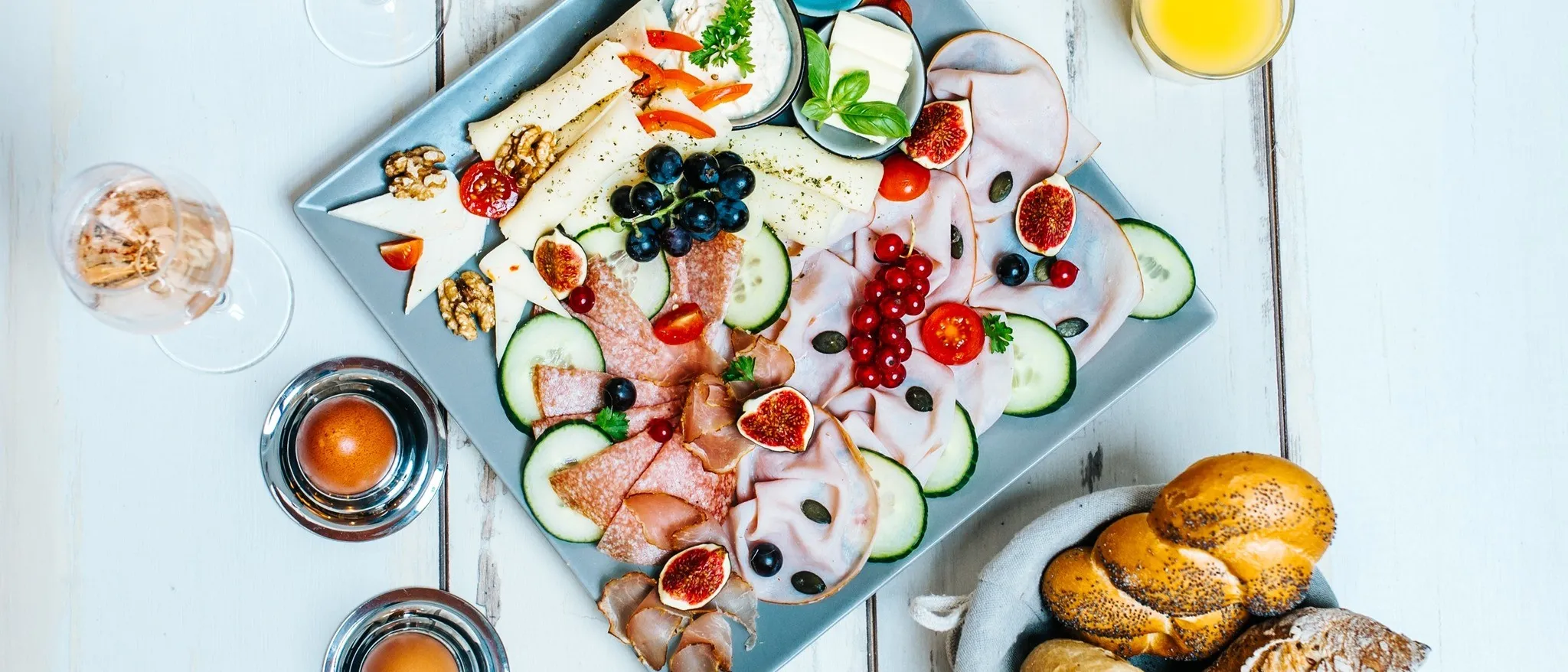 Ein blauer Teller mit verschiedenen Lebensmitteln wie Fleisch, Käse, Obst und Gemüse, begleitet von Brot, Eiern und Gläsern Orangensaft auf einem weißen Tisch.