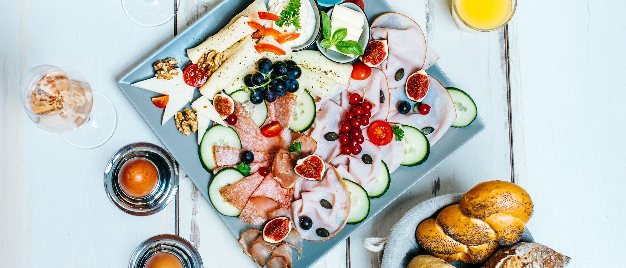 Ein blauer Teller mit verschiedenen Lebensmitteln wie Fleisch, Käse, Obst und Gemüse, begleitet von Brot, Eiern und Gläsern Orangensaft auf einem weißen Tisch.