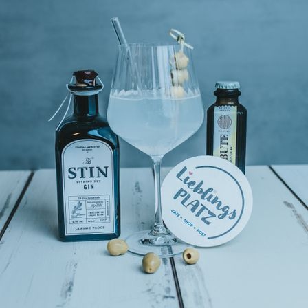 Ein Weinglas mit einem Cocktail steht auf einem Tisch mit einer Ginflasche, einer kleinen Flasche und einem Untersetzer.