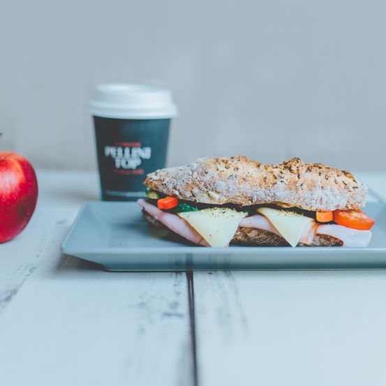 Ein Sandwich mit Schinken, Käse und Gemüse steht auf einem Teller neben einem Apfel und einer Tasse. Der Sandwich ist auf einem grauen Teller mit einer Tasse Kaffee im Hintergrund.