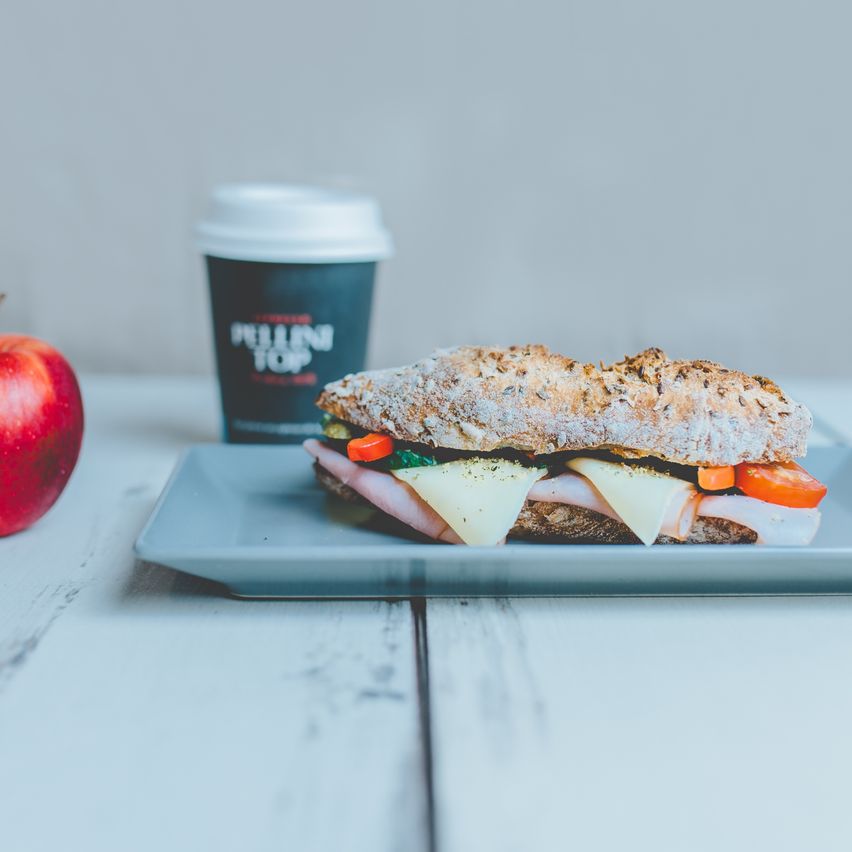 Ein Sandwich mit Schinken, Käse und Gemüse steht auf einem Teller neben einem Apfel und einer Tasse. Der Sandwich ist auf einem grauen Teller mit einer Tasse Kaffee im Hintergrund.