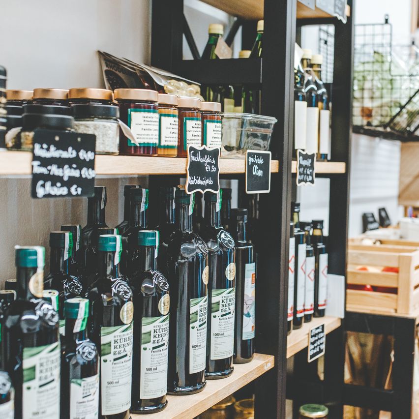 Ein Ladenregal zeigt mehrere Flaschen Olivenöl und andere Gewürze. Verschiedene Gläser sind beschriftet und auf oberen Regalen angeordnet.