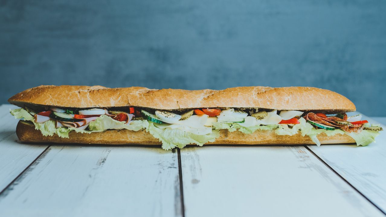 Ein langes Sandwich mit verschiedenen Zutaten wie Salat, Tomaten und Käse auf einem weißen Tisch.