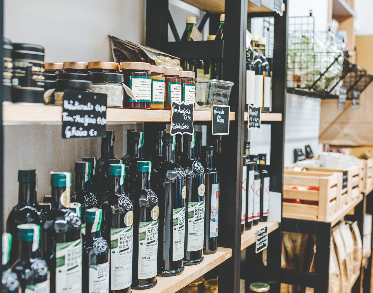 Ein Laden mit Regalen voller Flaschen und Gläser. Verschiedene Produkte wie Öle, Marmeladen und Gewürze sind ausgestellt. Die Regale sind mit Etiketten und Preisschildern organisiert. Ein Holzkiste ist im Hintergrund sichtbar.