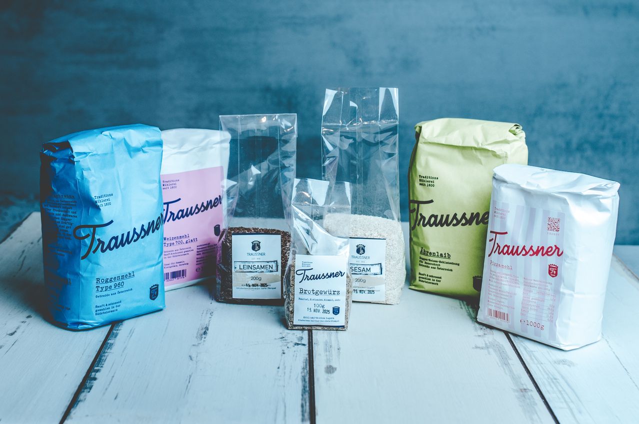Verschiedene transparente und farbige Tüten mit Mehl und Samen, gekennzeichnet mit 'Traussner', sind auf einer weißen Holzoberfläche ausgestellt. Die Tüten enthalten verschiedene Arten von Mehl und Samen, einige mit Verfallsdaten.
