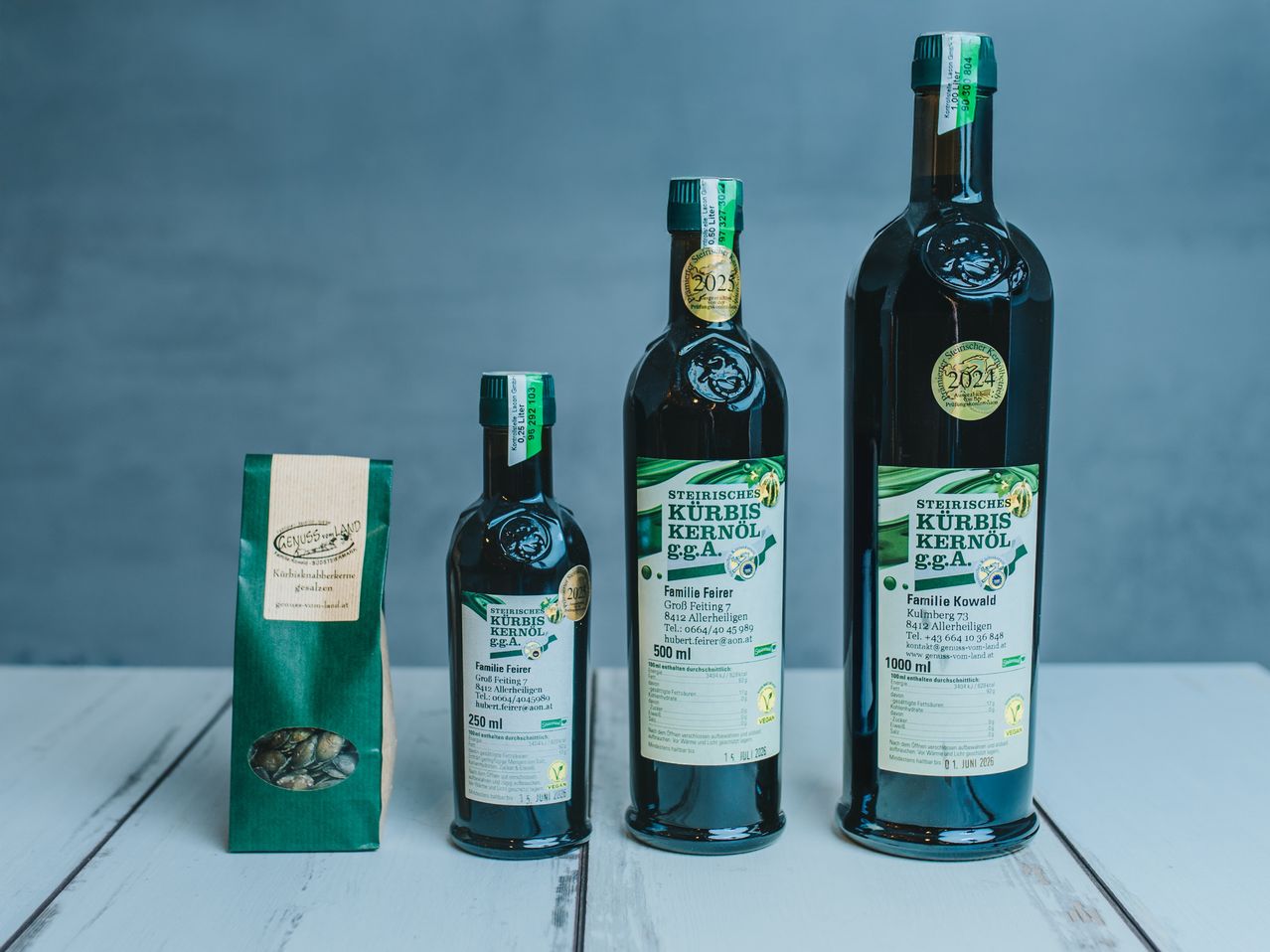 Drei Flaschen mit grünen Etiketten von 'Steirisches Kurbiskernöl' sind auf einer weißen Oberfläche angeordnet. Ein grüner Beutel mit Etikett ist links neben den Flaschen platziert. Die Flaschen haben grüne und goldene Etiketten, wobei eine 'Famili Fairer', eine andere 'Familie Kowald' und die dritte 'Steirisches Kurbiskernöl g.g.A.' mit dem Jahr 2024 beschriftet ist.