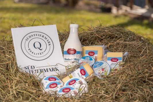 Milchprodukte einschließlich einer Milchflasche und verschiedener Käsesorten auf einem Heubett mit einem Südsteiermark-Qualitätszeichen im Hintergrund.