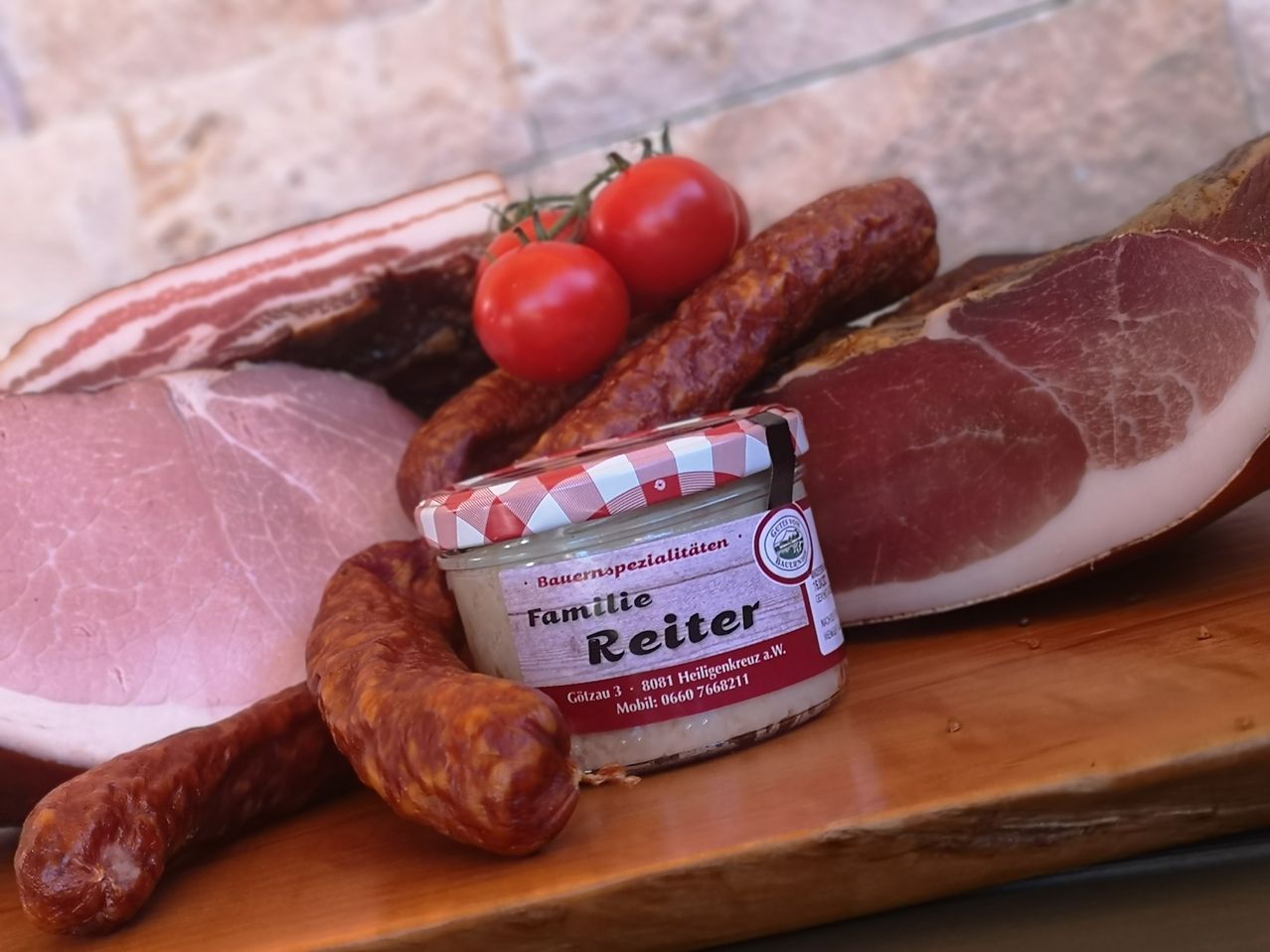 Auf einem Holzbrett sind verschiedene Fleischsorten zu sehen, darunter Würste, Speck und ein Glas Famie Reiter. Drei Tomaten sind ebenfalls auf dem Brett vorhanden.
