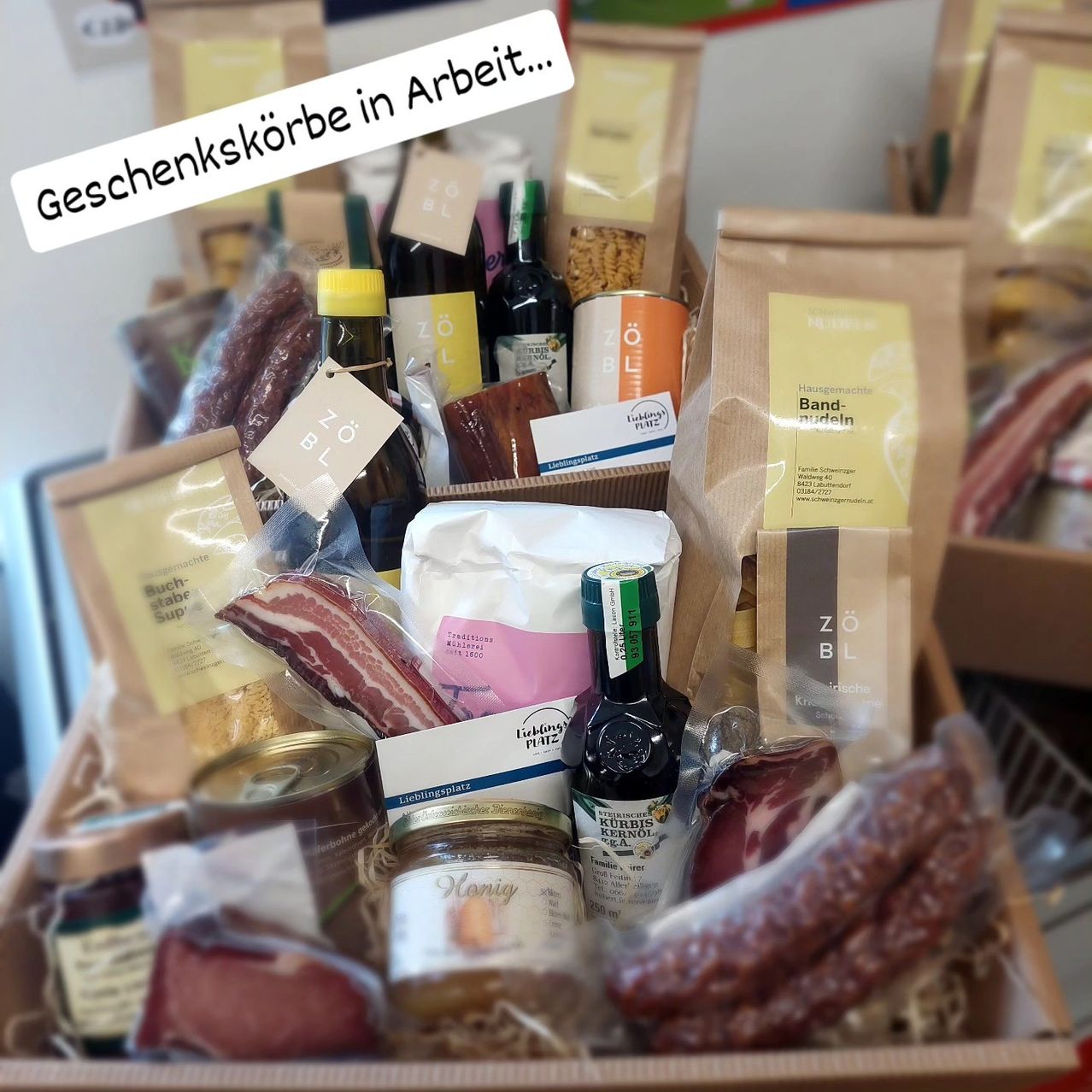 Eine Schachtel mit verschiedenen Lebensmitteln wie Speck, Wein, Honig und Nudeln. Einige Artikel sind in Plastiktüten, andere in Gläsern und Flaschen. Die Schachtel hat ein Etikett mit der Aufschrift 'Geschenkkörbe in Arbeit...'.