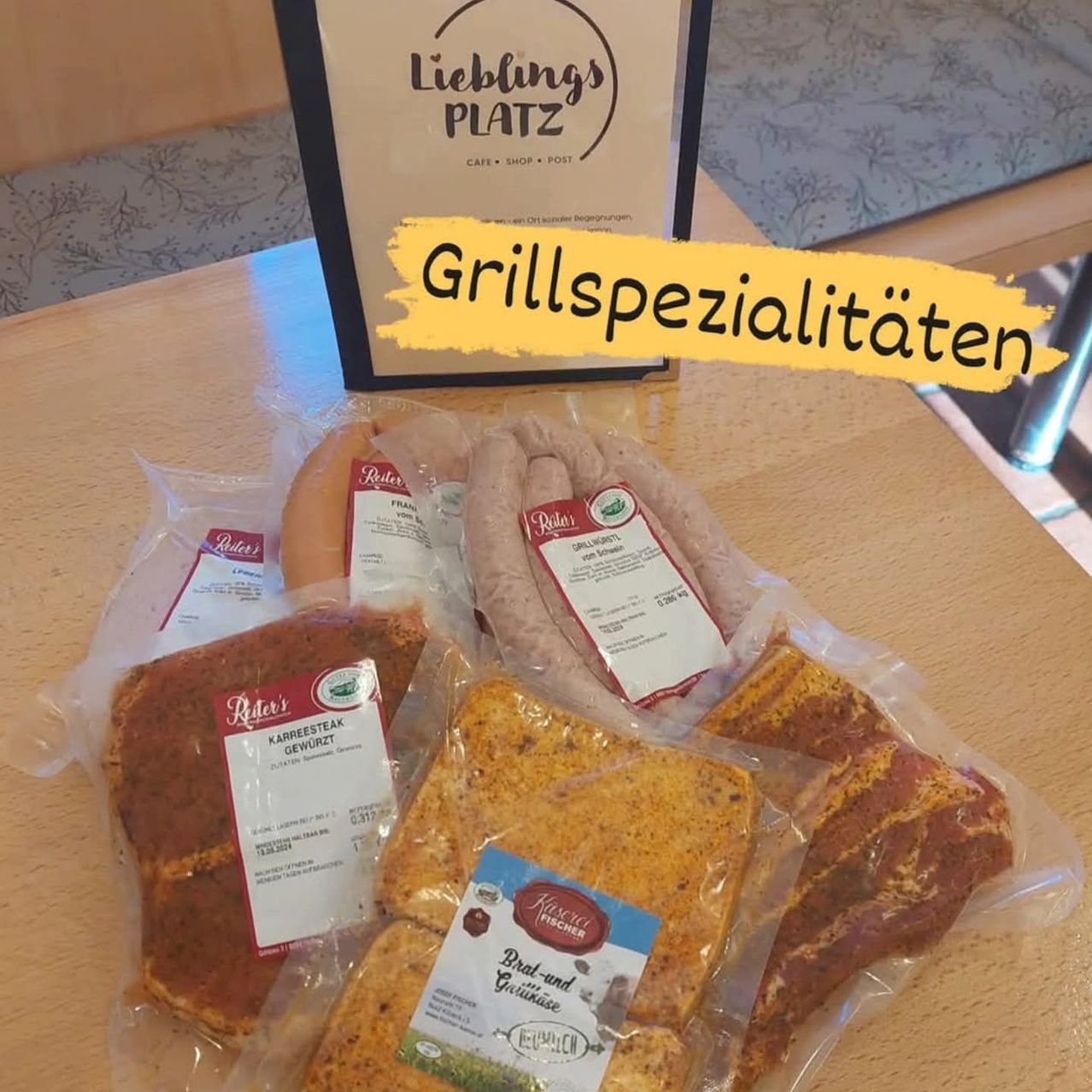 Auf einem Holztisch sind verschiedene Fleischsorten in durchsichtigen Plastiktüten zu sehen, darunter Würste und ein Stück mit der Aufschrift 'Karreesteak Gewurzt'. Darüber befindet sich ein Schild für 'Lieblings PLATZ' mit dem Text 'Grillsspezialitat'.