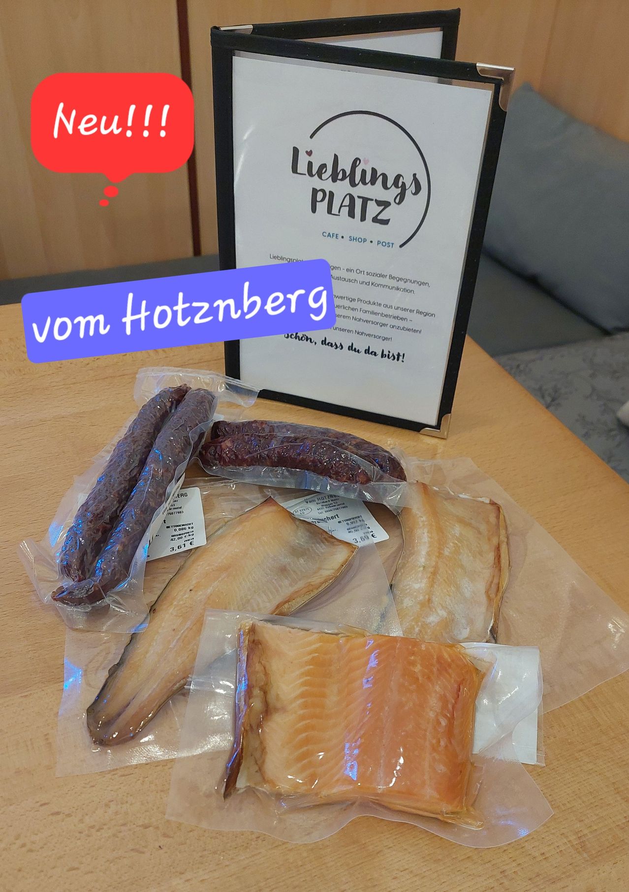 Verpackte Fleisch- und Fischprodukte sind auf einem Tisch ausgestellt, auf dem ein Schild mit der Aufschrift 'Lieblingsplatz' steht. Die Artikel umfassen Würste und Lachsscheiben.