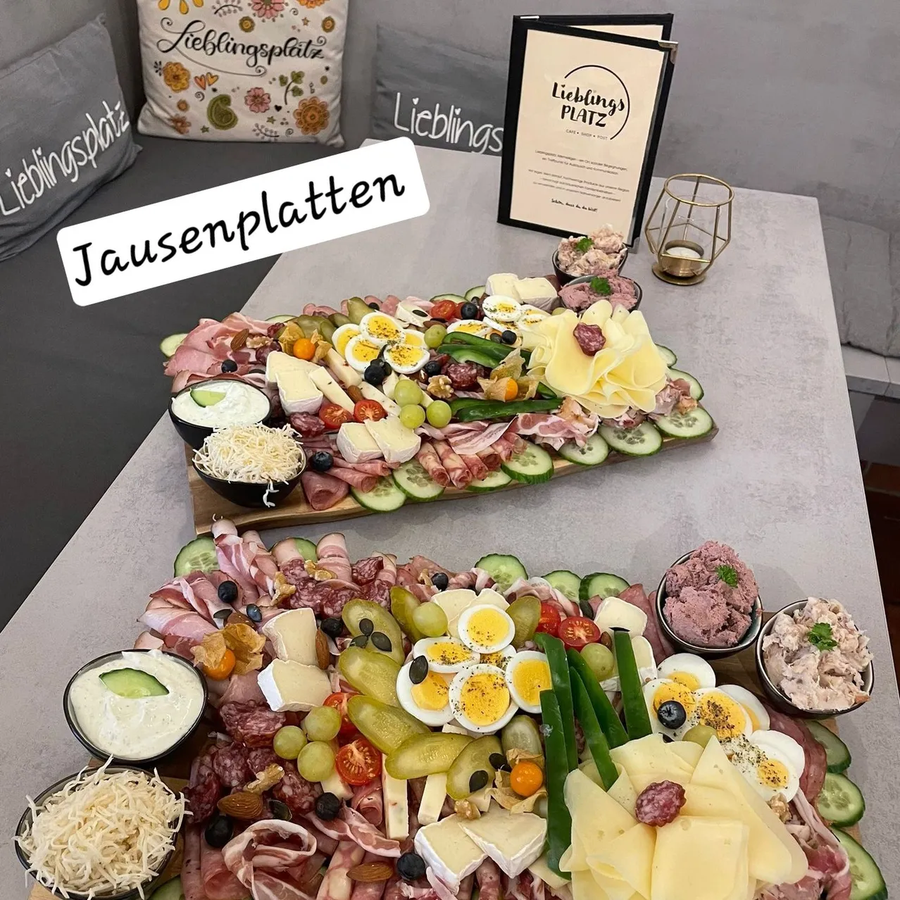 Ein Tisch mit zwei großen Platten mit Charcuterie, die eine Vielzahl von Fleischsorten, Käse, Gemüse und Aufstrichen zeigen. Die obere Platte enthält geschnittenes Fleisch, Käse, Eier und Gurken. Die untere Platte hat eine Blumenarrangement aus Käse, Oliven und Gemüse. Ein Schild mit dem Text 'Jausenplätten' steht auf dem Tisch.