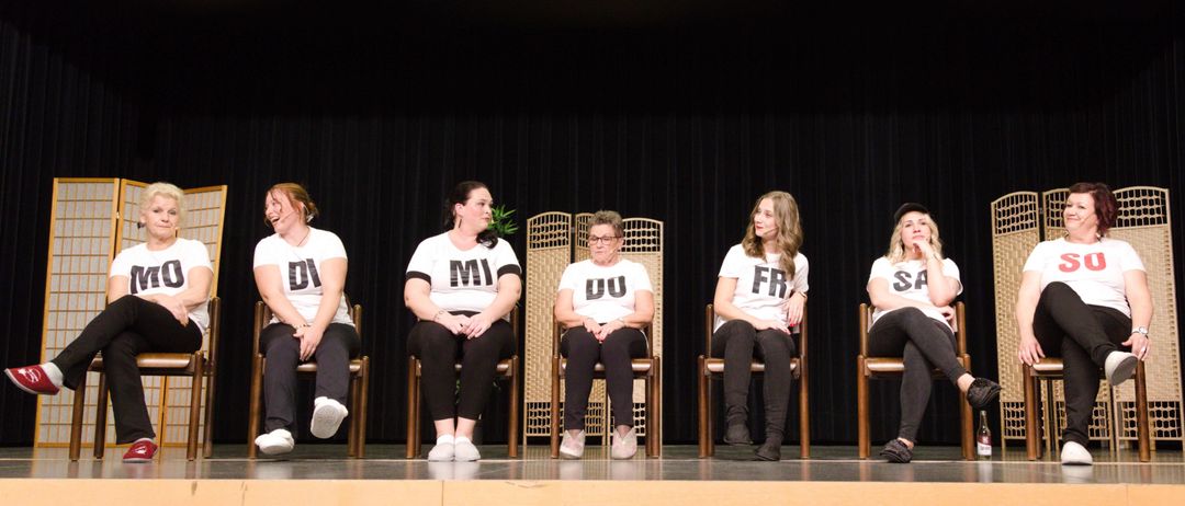 Fünf Frauen sitzen auf Stühlern auf einer Bühne, wahrscheinlich während einer Vorstellung. Jede hat einen anderen Anfangsbuchstaben auf ihrem Shirt, wie DI, MI, OU, FR und SA.