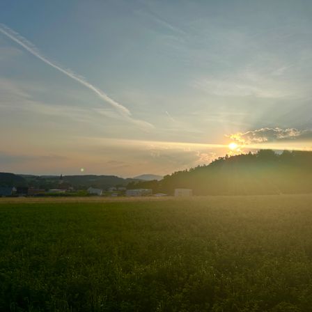 Sonnenuntergang über einer ländlichen Landschaft mit einem Feld, Bäumen und einem kleinen Dorf in der Ferne. Die Sonne geht hinter den Wolken unter und wirft ein warmes Licht.